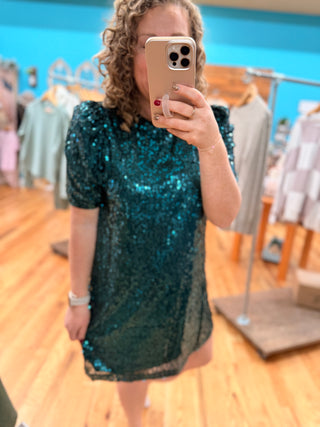 Person wearing a teal sequin dress taking a mirror selfie in a clothing store.